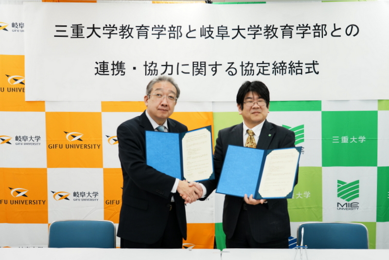 協定書への署名後、手を取り合う山田雅博岐阜大学教育学部長(左)と伊藤信成三重大学教育学部長(右)